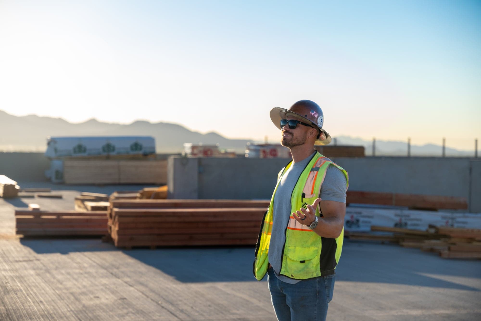 Safirst safety consultant in hi-vis vest and hard hat on an elevated deck at sunrise, lumber stacked behind, desert mountains on the horizon