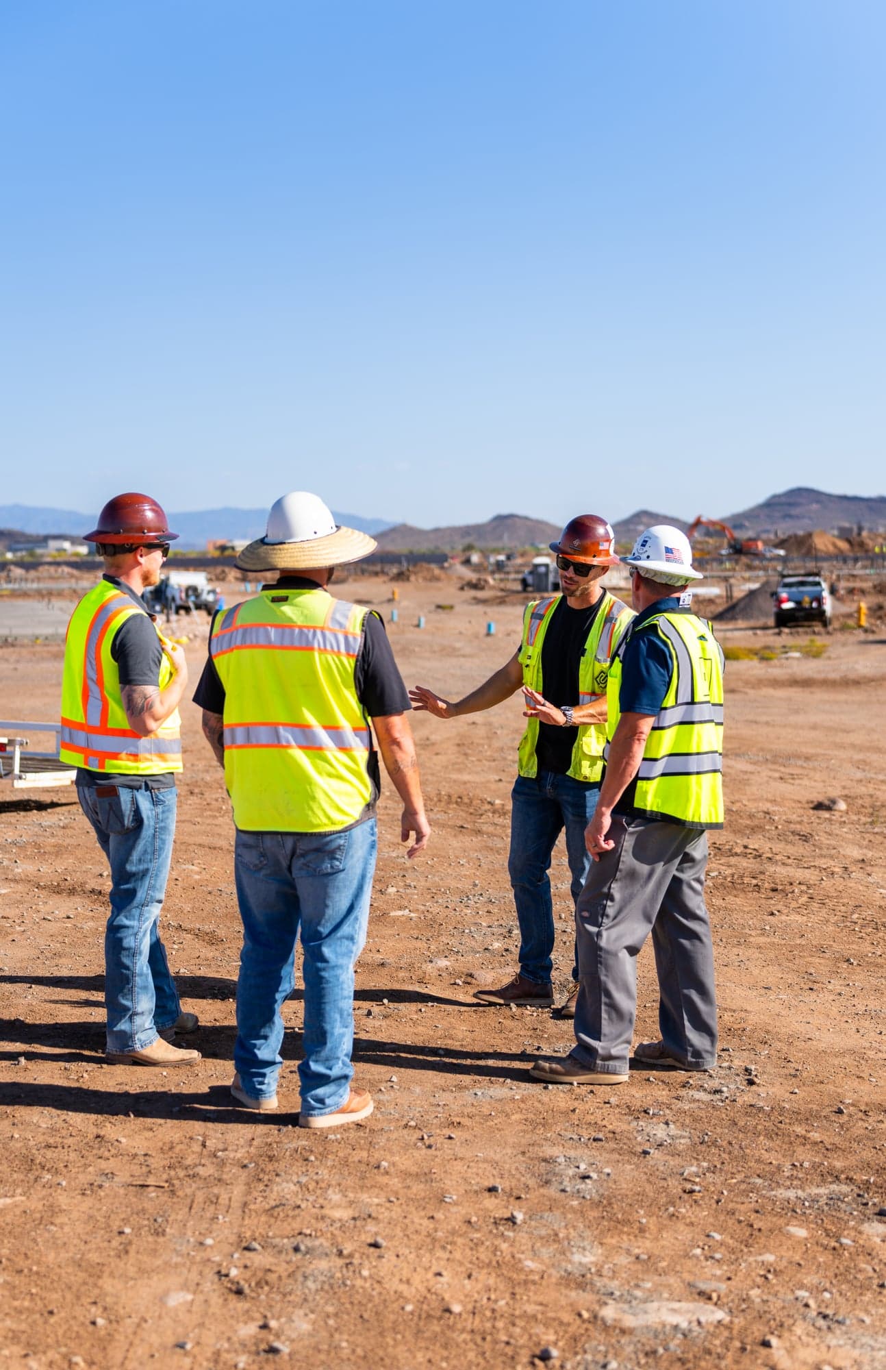 Four workers in hi-vis vests and hard hats gathered in a circle for a pre-task safety briefing on a graded site