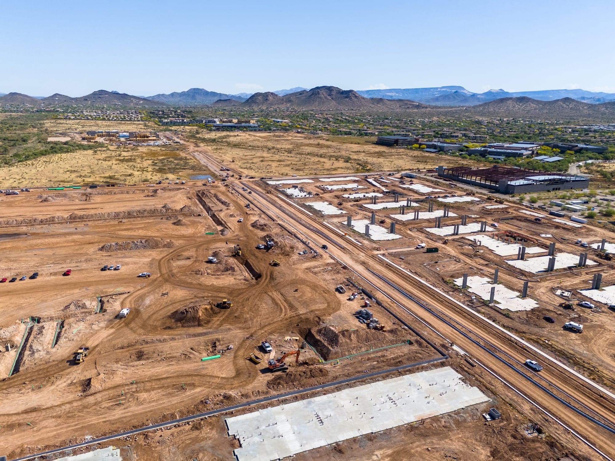 Aerial view over a large Arizona mass-grading site with fresh concrete pads, haul roads and mountains on the horizon