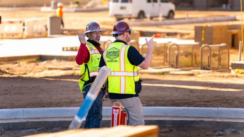 A Safirst rep gesturing with both hands as he talks to a worker on a graded pad, curb and formwork behind them
