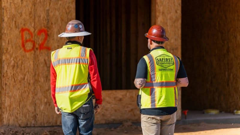 Two Safirst staff, one in a wide-brim sun hard hat, standing at an OSB-sheathed wall spray-marked 'B2'