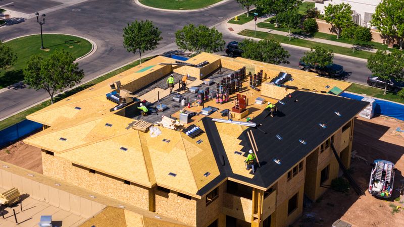 Aerial view of a roofing crew working on a sheathed two-story building, materials staged across the roof deck