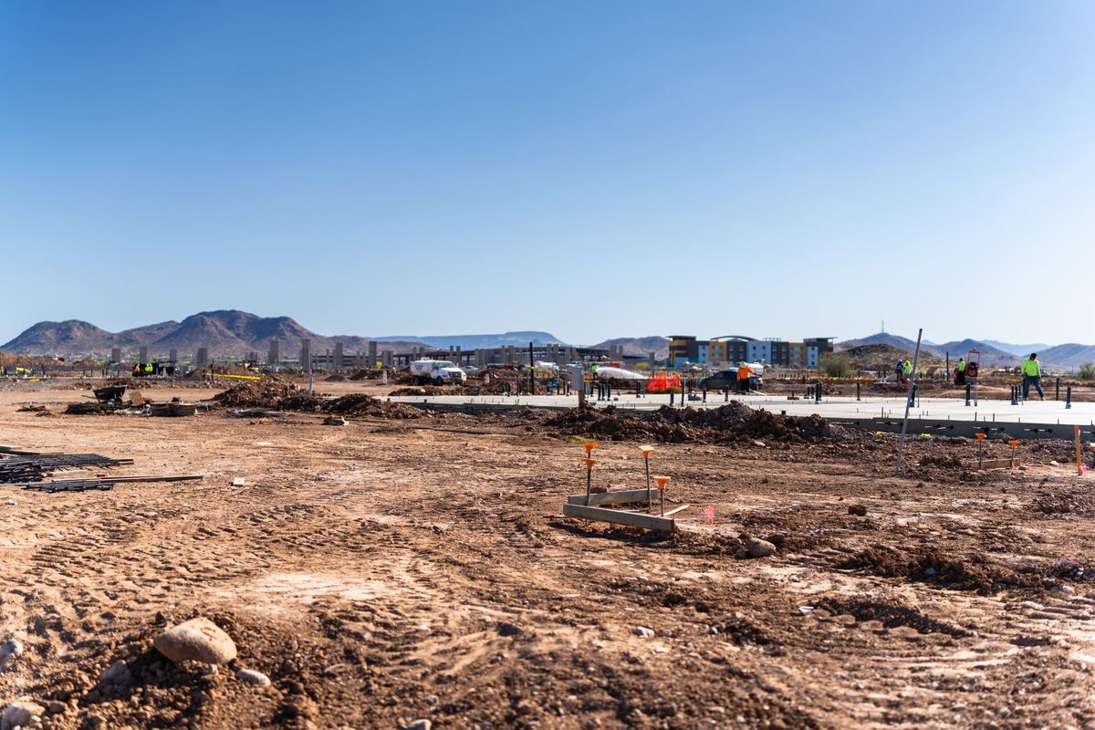 Zapata de concreto sobre un terreno nivelado con sus varillas verticales cubiertas con tapas protectoras anaranjadas, y una cuadrilla trabajando al fondo