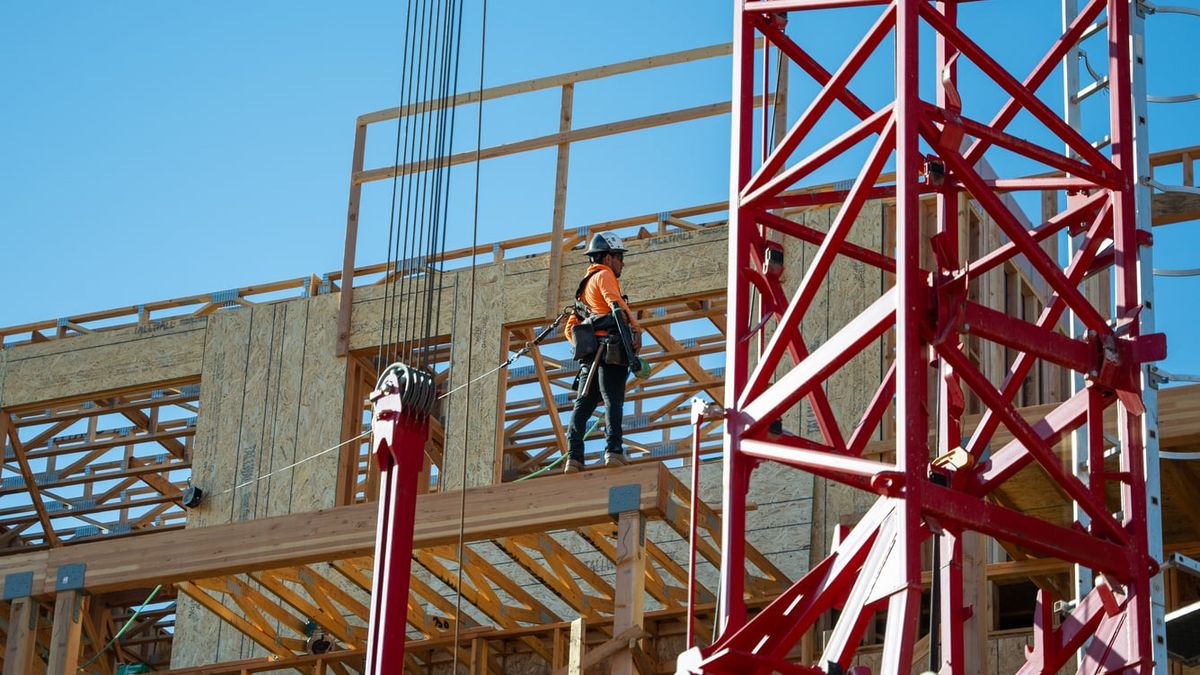 Worker in a hard hat and full-body harness, lanyard attached, standing on the open upper floor of a wood-framed building beside a tower crane mast