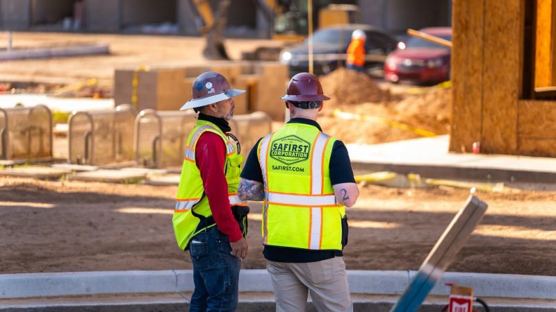 A Safirst rep and a foreman in a red shirt standing face to face on a graded pad, both in hi-vis and brim hard hats